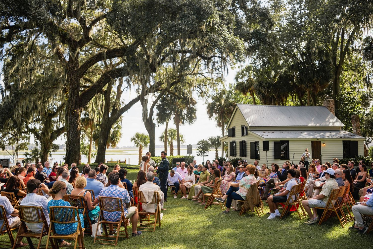 A large outdoor gathering of people seated in chairs under shady oak trees during a daytime event.