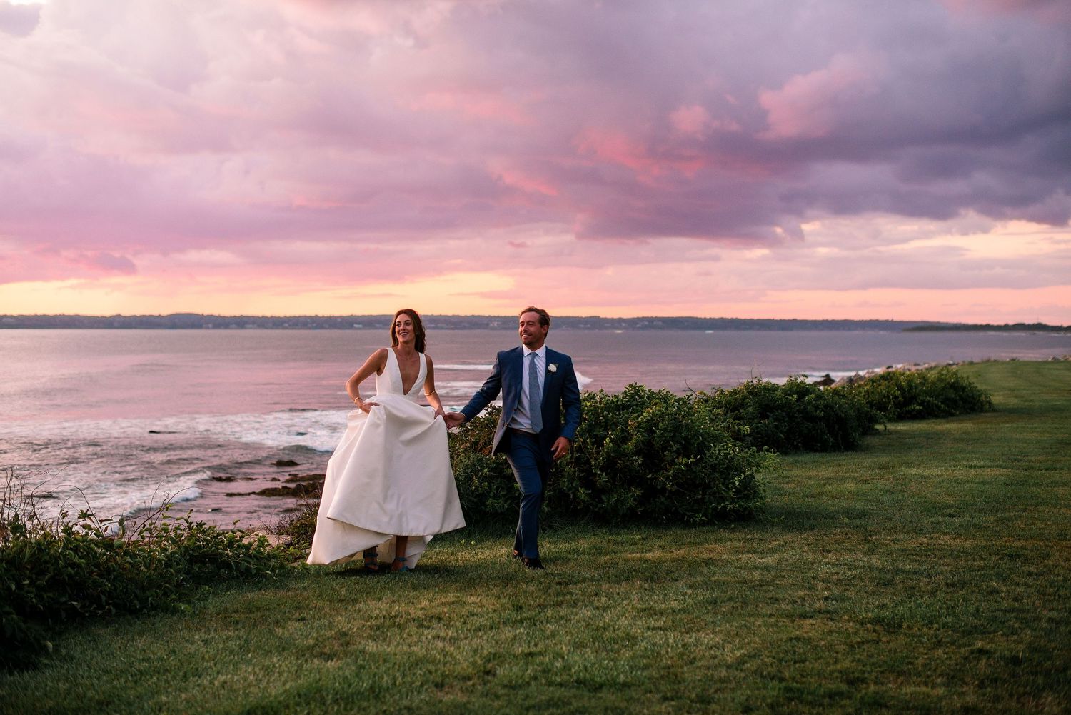 A couple in wedding attire walks hand in hand along a grassy coastal path during a vibrant pink sunset.