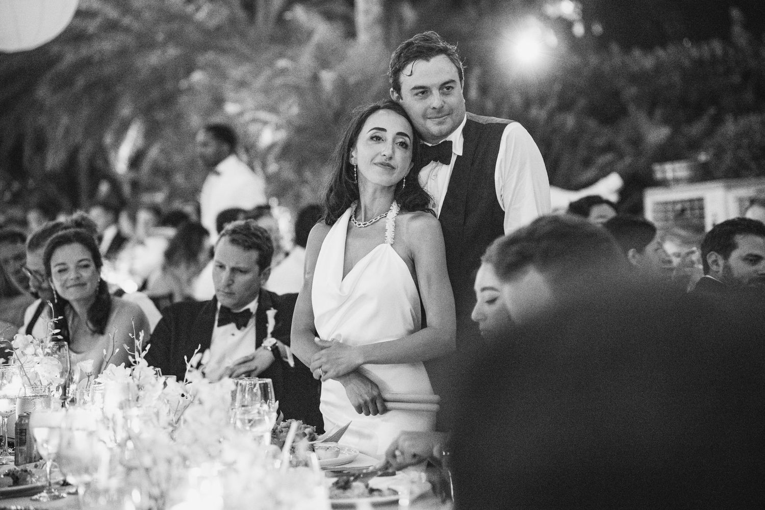 A black and white wedding reception photo showing a couple embracing at their candlelit dinner table.