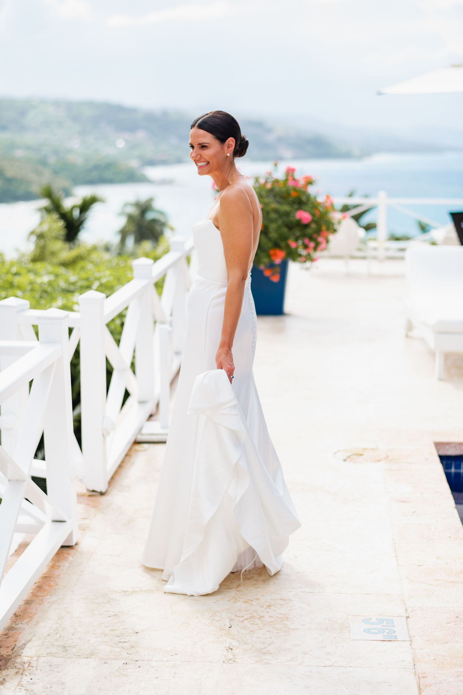 A bride in a flowing white wedding dress twirls on a seaside terrace overlooking tropical waters and mountains.