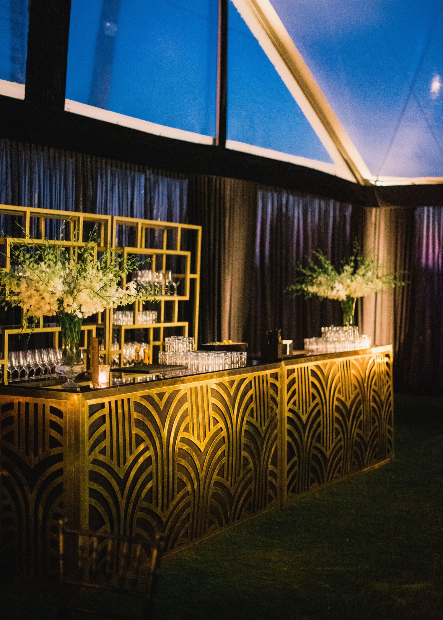 Art deco-style bar with gold geometric patterns and floral centerpieces illuminated under dramatic lighting at an elegant event.