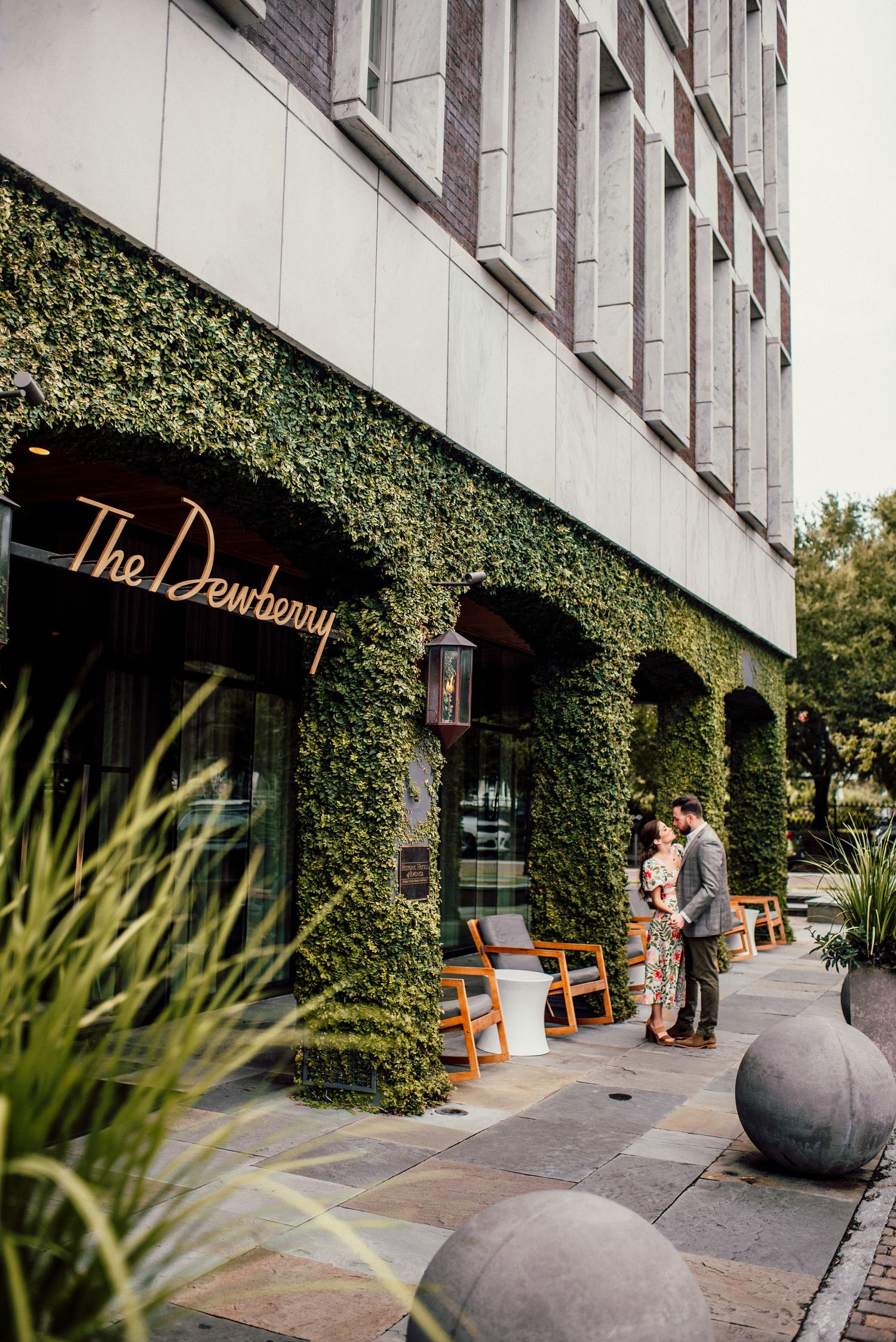 A modern building exterior with green ivy walls, concrete spheres, and orange chairs along the sidewalk.