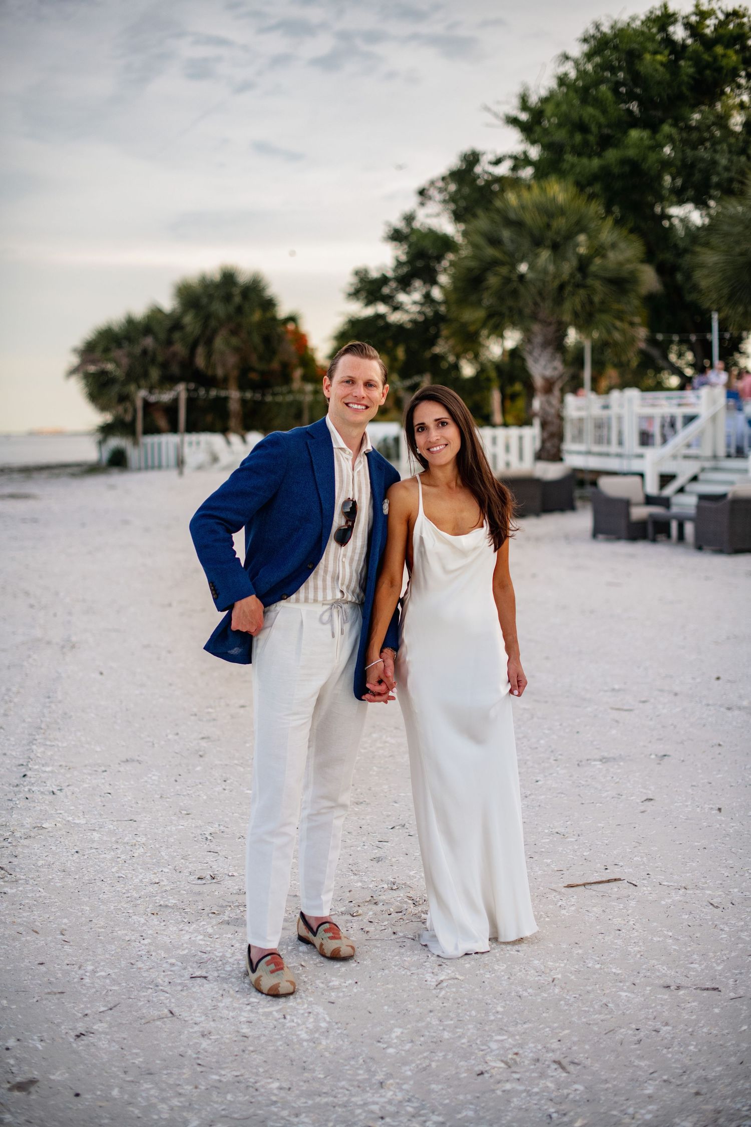 A couple in coordinating blue and white outfits poses playfully on a sandy beach at sunset.