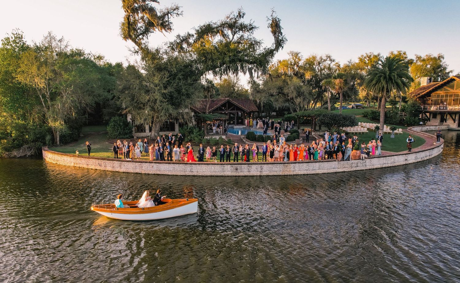Panoramic view of boats and guests gathered along a scenic lakeside park during a sunset celebration.