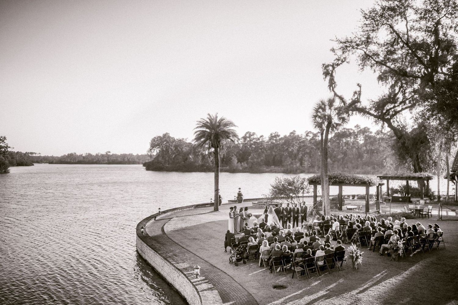 Panoramic view of a waterfront wedding ceremony setup with white chairs and palm trees along a scenic shoreline.