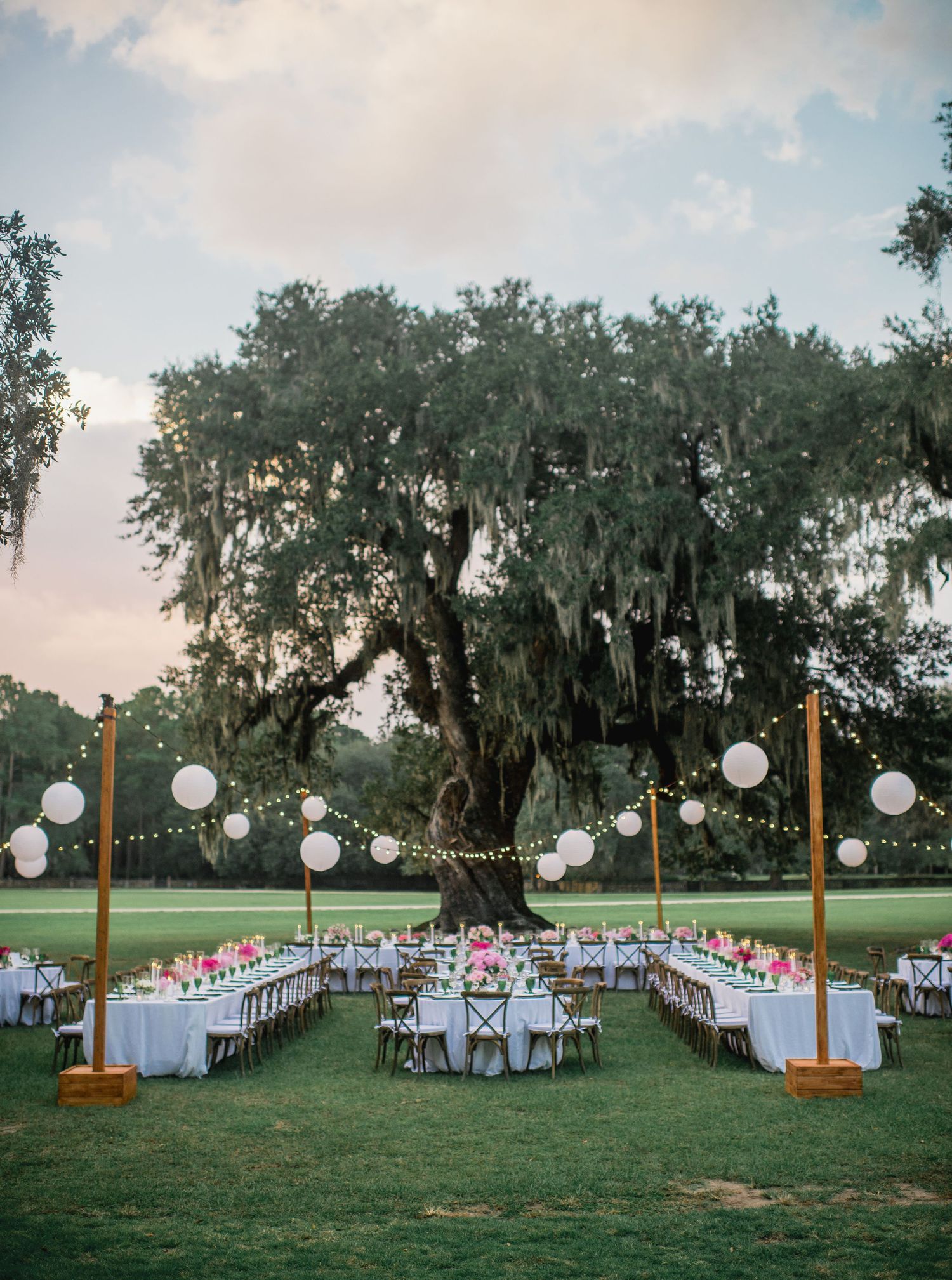 Elegant outdoor wedding reception setup with long tables and hanging lanterns under Spanish moss-draped trees at dusk.