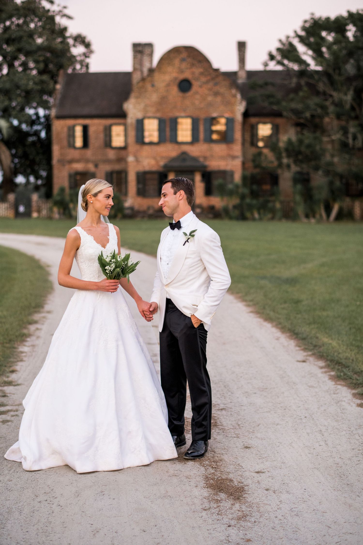 A couple in wedding attire shares romantic moments on a gravel path with a brick building in the background.