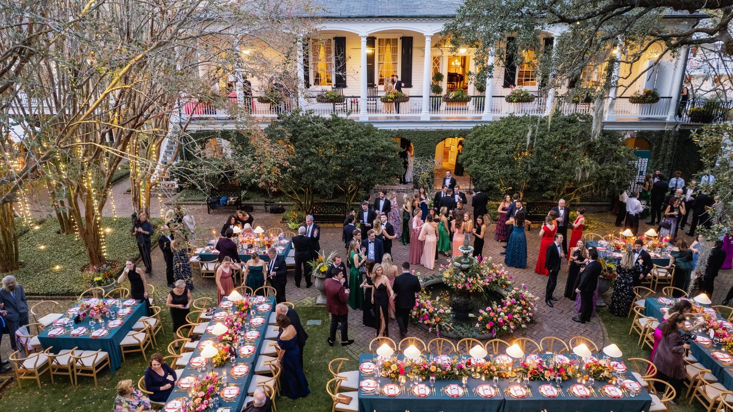 Outdoor wedding reception setup with long tables and string lights glowing at dusk in a garden setting.