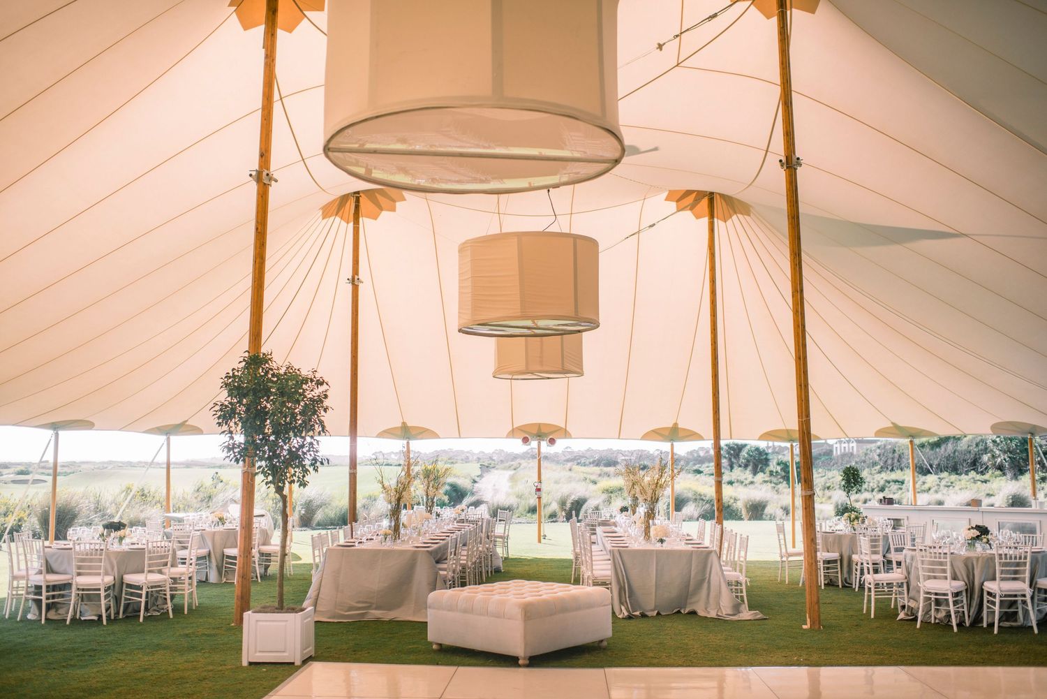 Interior of an elegant white wedding tent with round tables, chandeliers, and pole draping for a luxurious reception setup.