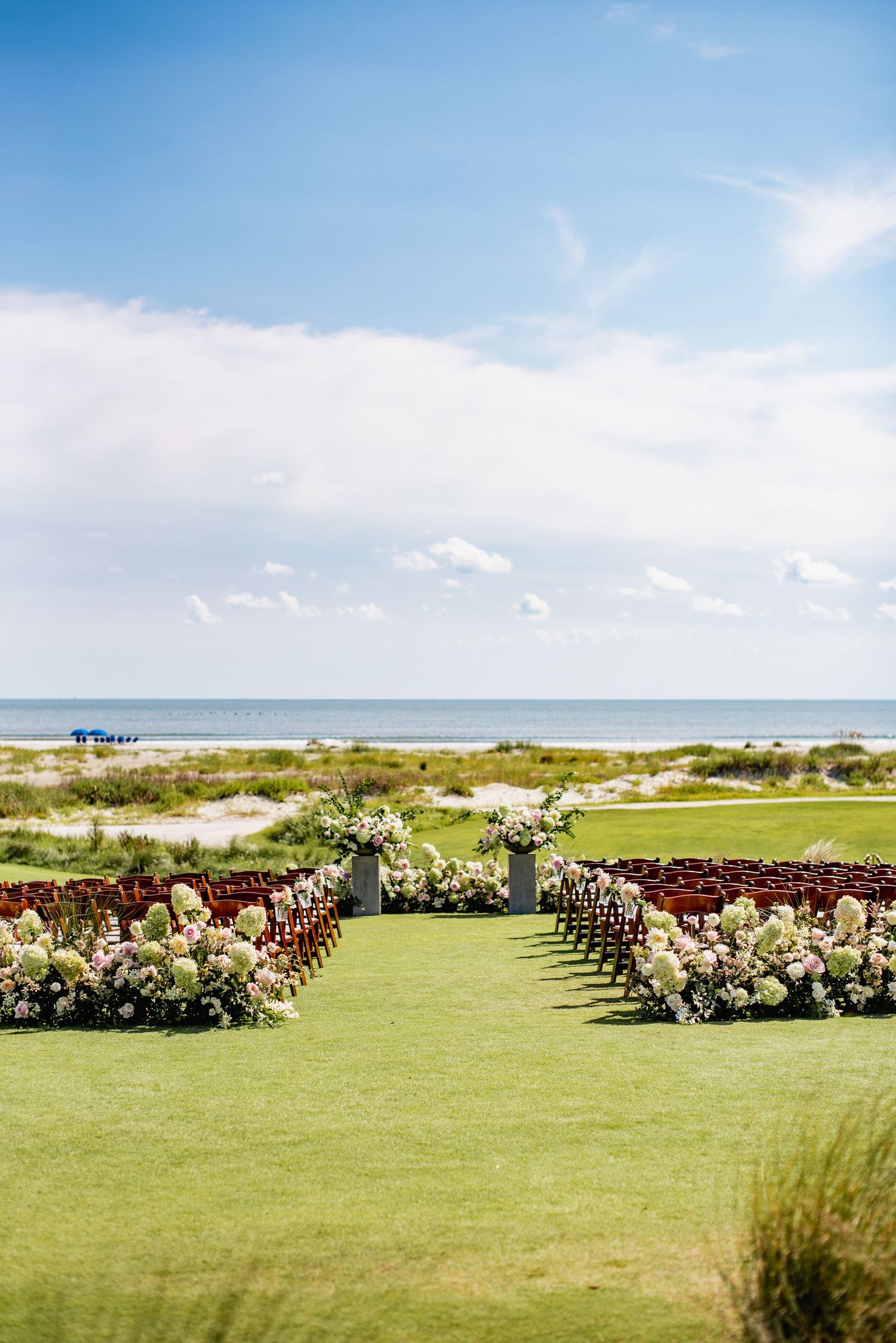 Rows of wooden chairs arranged on a lush green lawn overlooking a pristine beach and ocean on a sunny day.