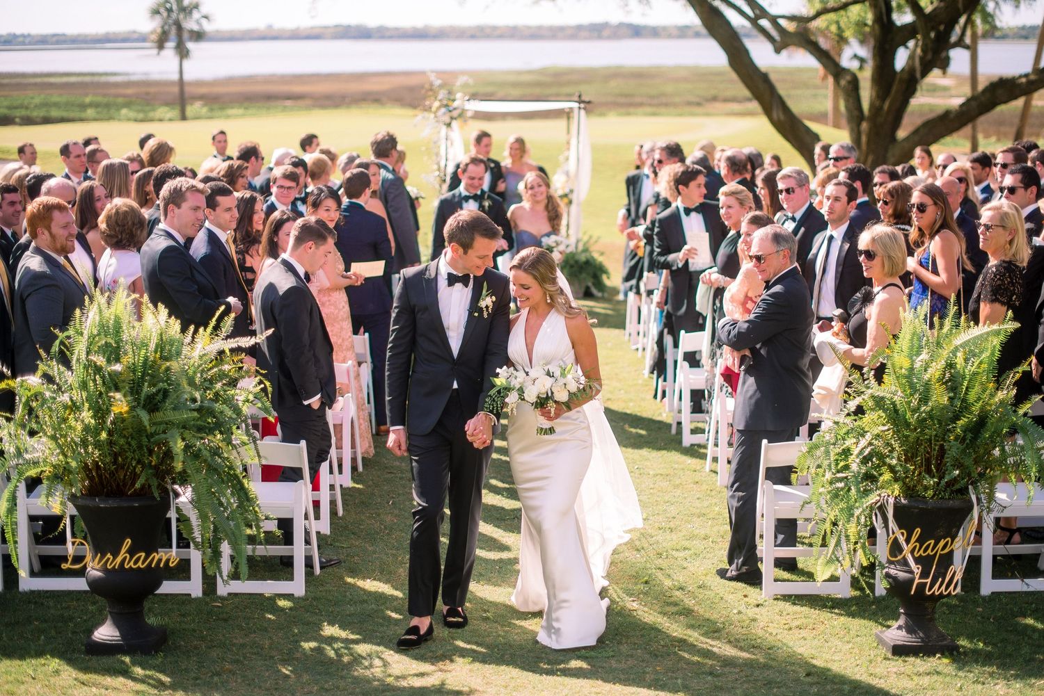 A wedding ceremony takes place outdoors on a lush green lawn with palm trees and water views in the background.