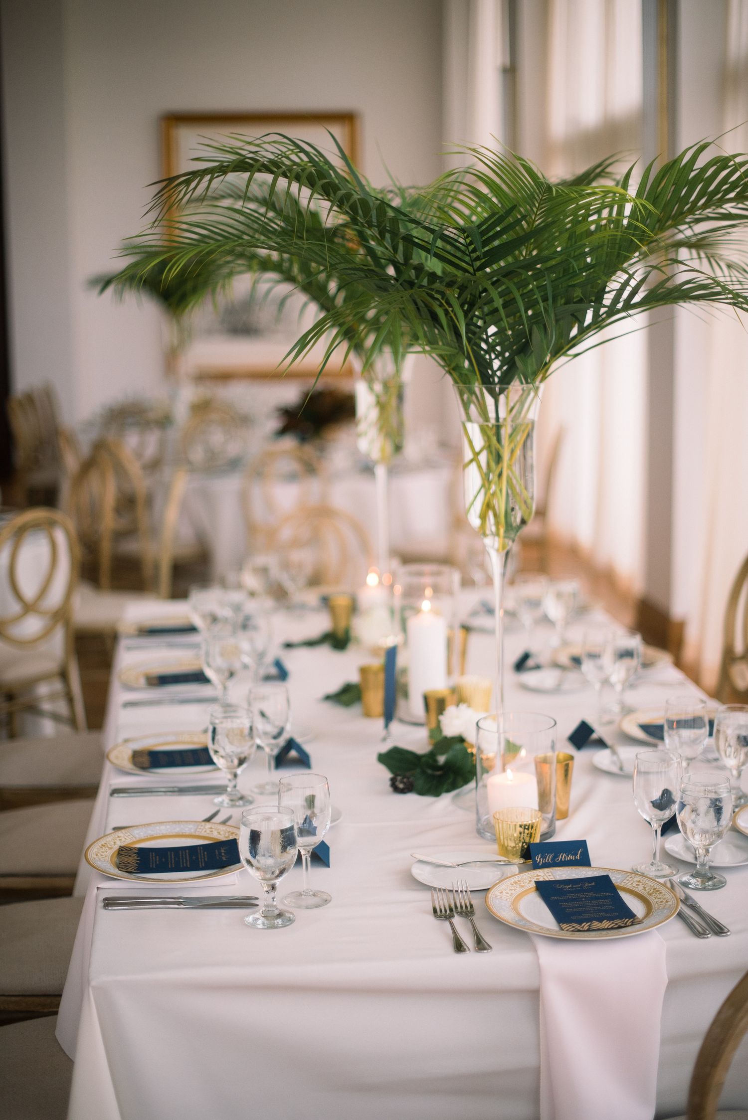 Elegant wedding reception tables decorated with tropical palm fronds, white linens, and navy blue place settings.