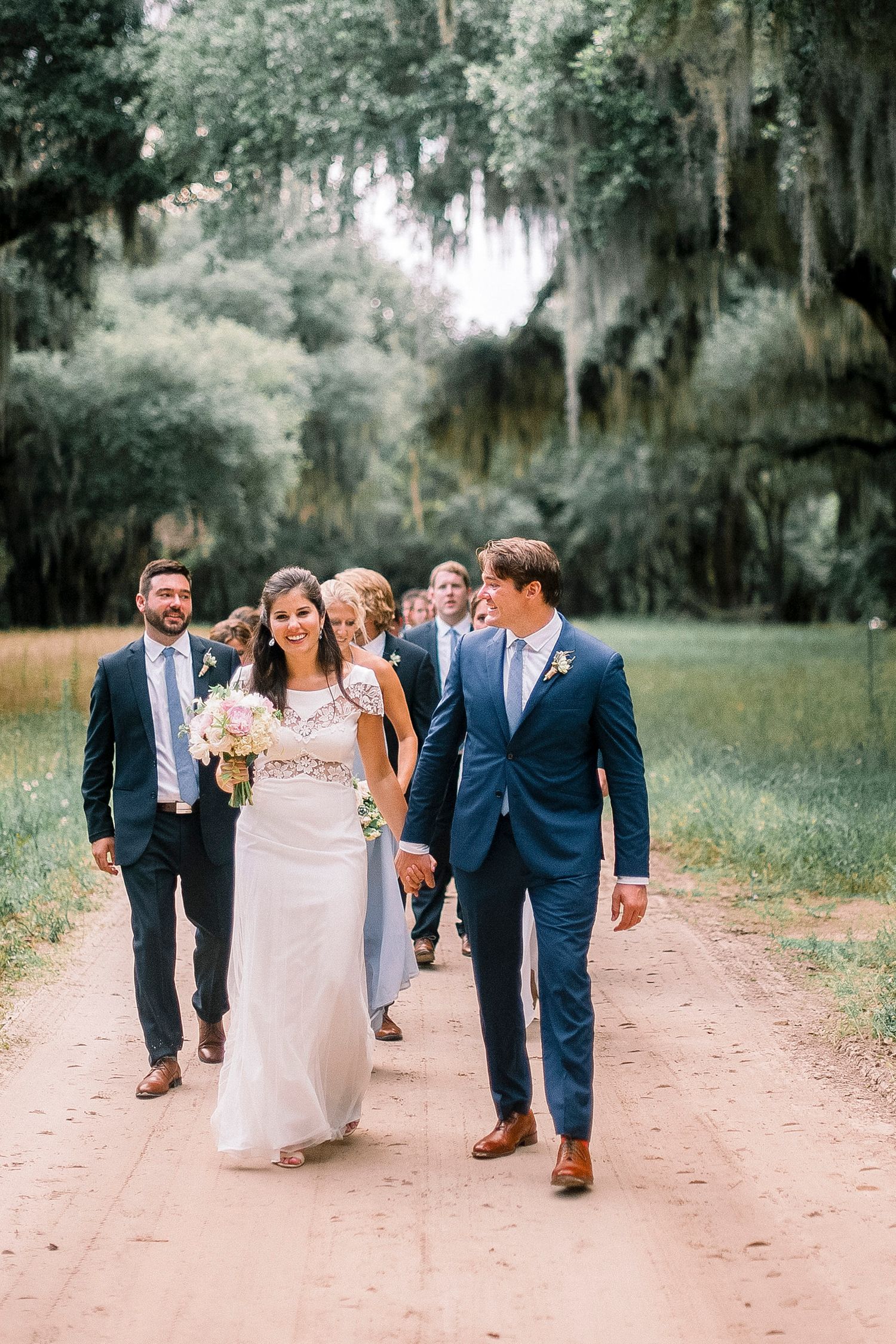 Wedding party walking together on tree-lined dirt path in formal attire.