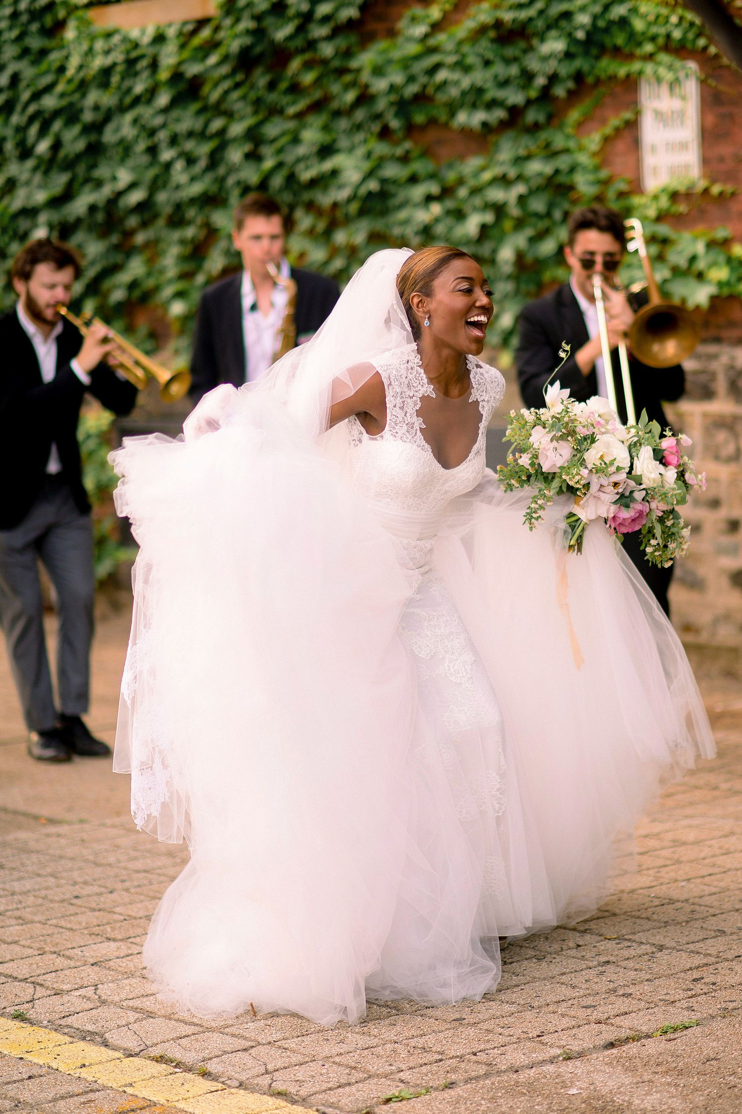 Bridesmaids in flowing white dresses twirl and dance on stone pavement surrounded by green ivy.