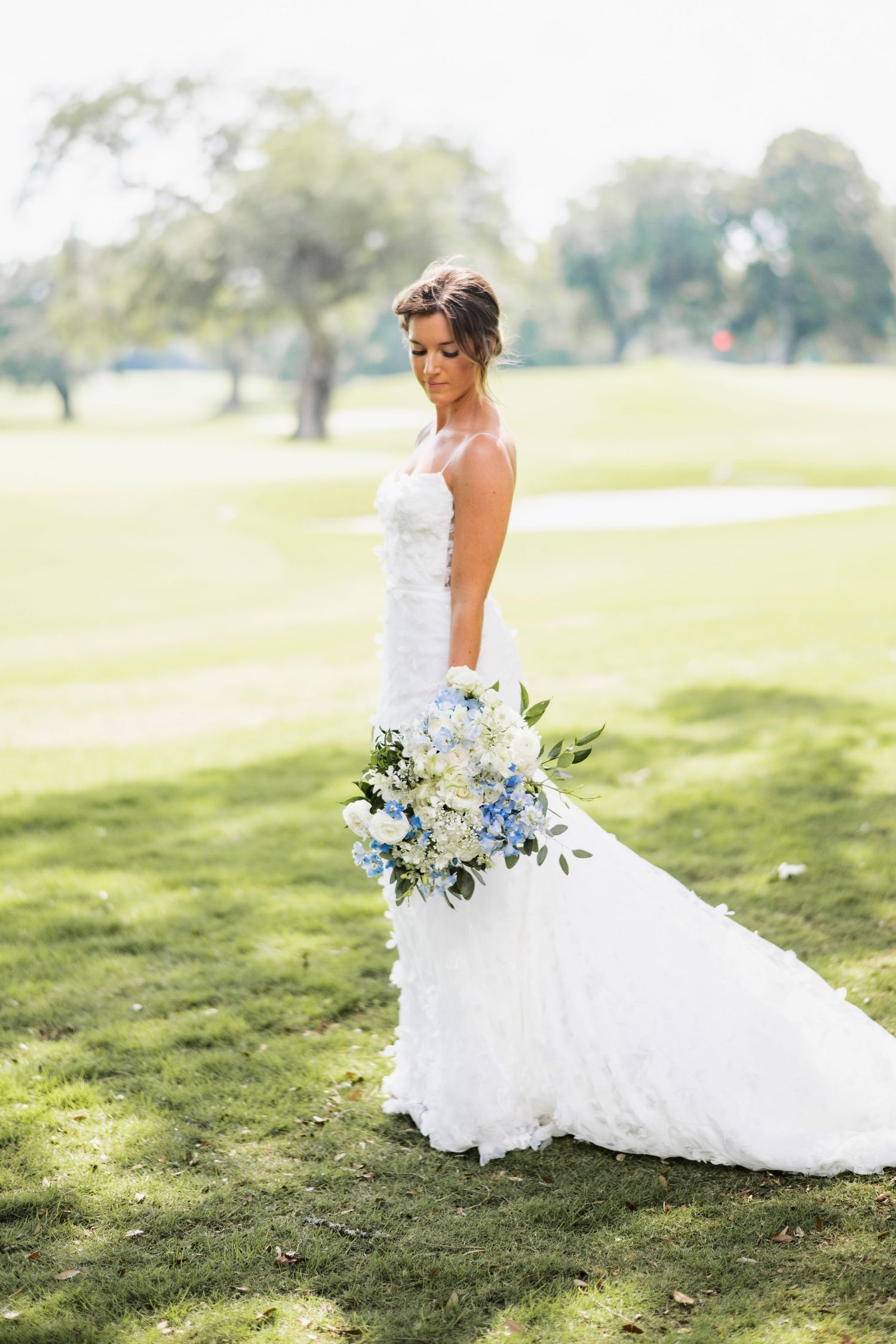 A white wedding gown with a flowing train and blue bouquet photographed on a sunlit golf course.
