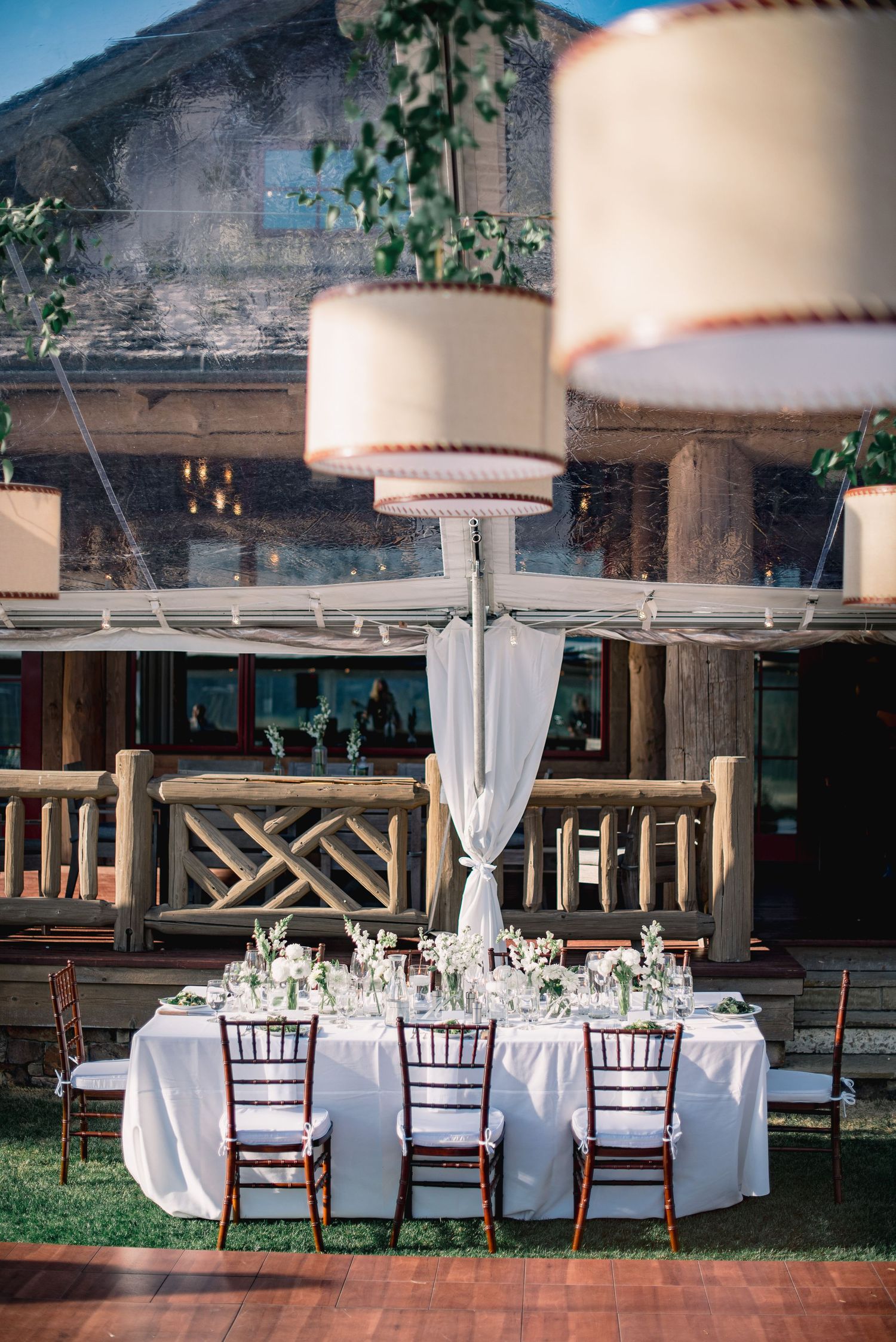 Elegant outdoor wedding reception setup with white linens, mahogany chairs and dramatic drum shade pendant lighting.