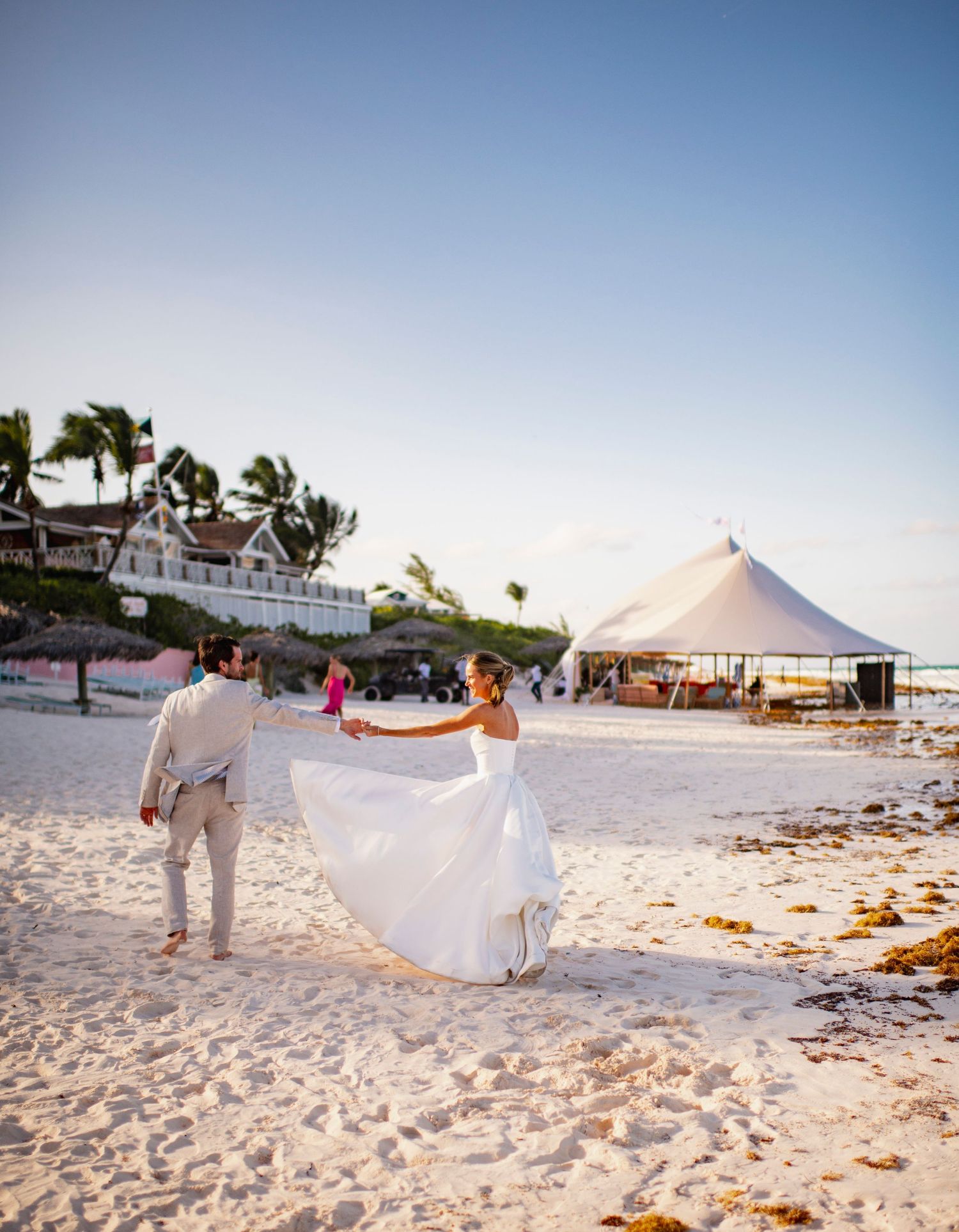 Couple walking on a beach at sunset with flowing white dress and beach tent reception setup visible.