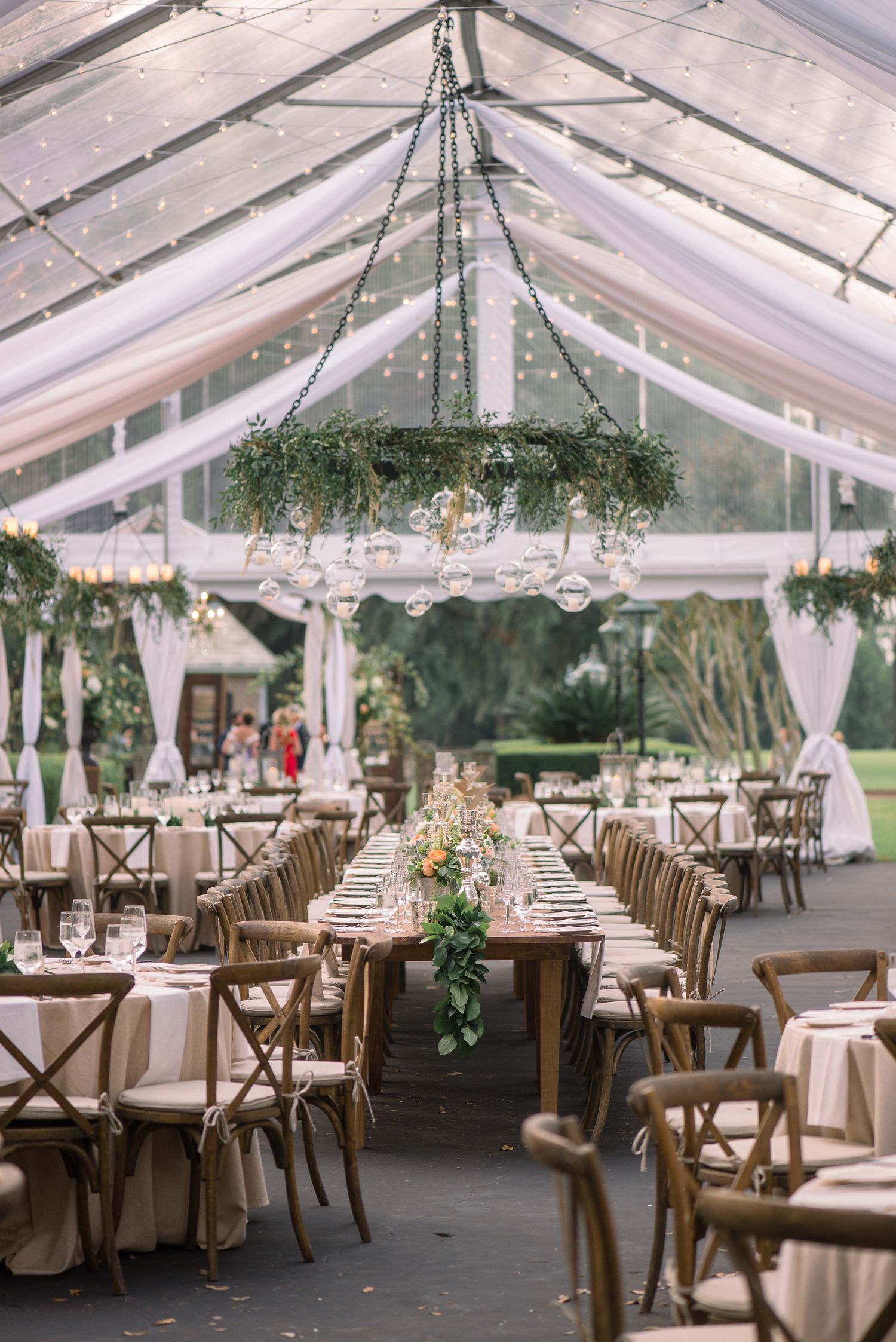 Elegant wedding reception setup in a clear tent with wooden cross-back chairs, white linens, and hanging greenery decor.