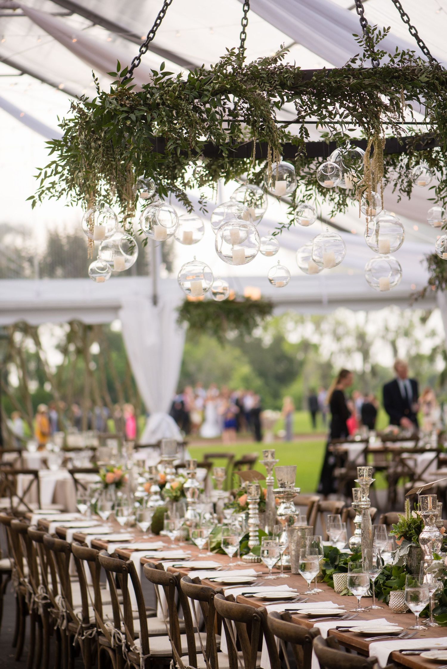 Elegant wedding reception table settings under a white tent with hanging greenery and crystal decorations.