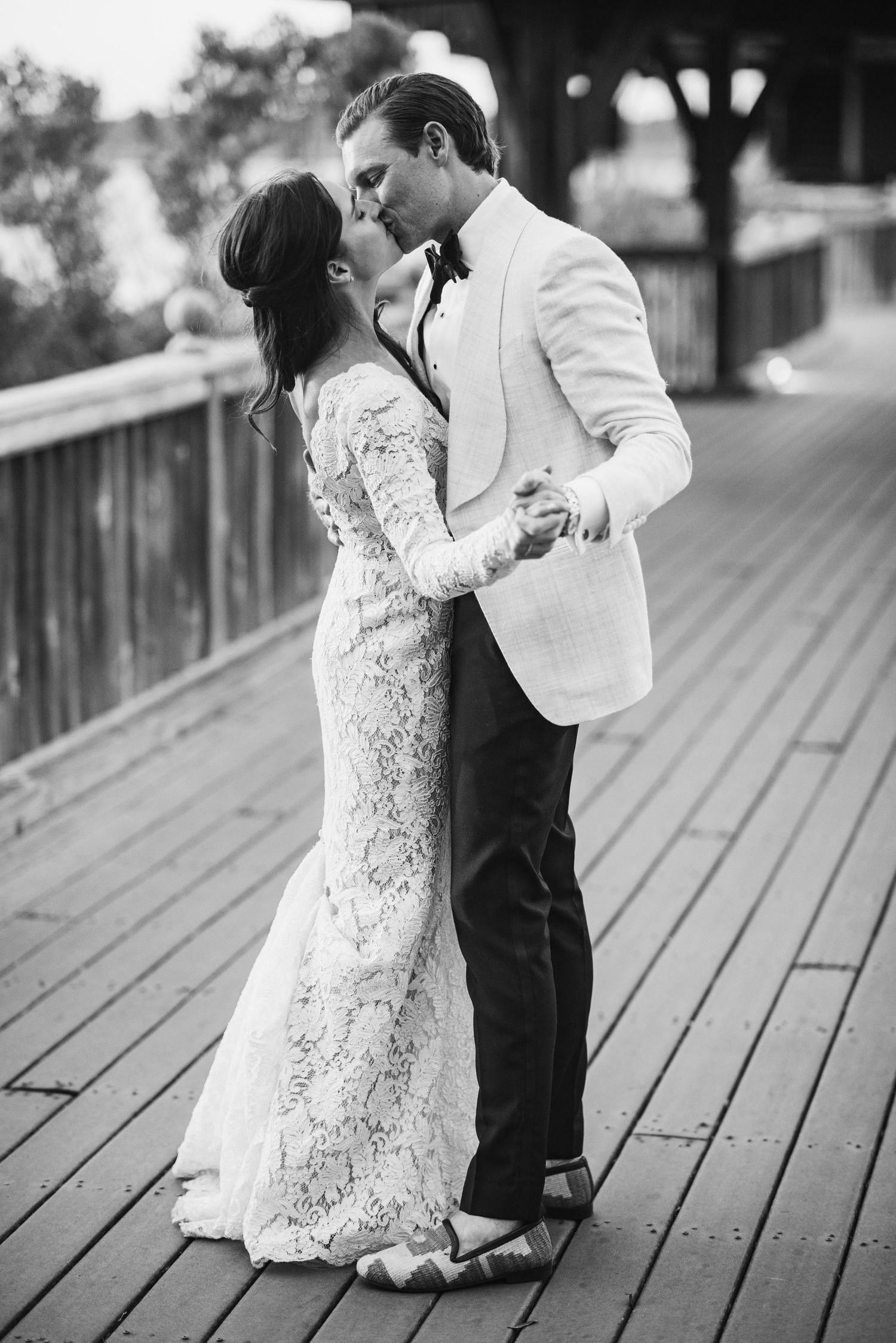 A couple in wedding attire share an intimate dance moment on a wooden deck in black and white photography.