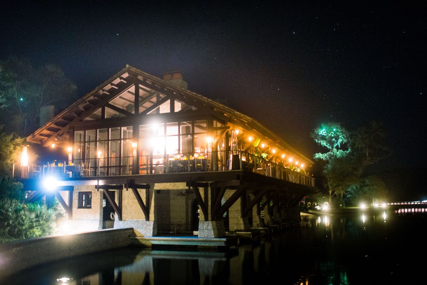 Illuminated wooden lake house with glass walls glows warmly at night against dark sky and reflective water.