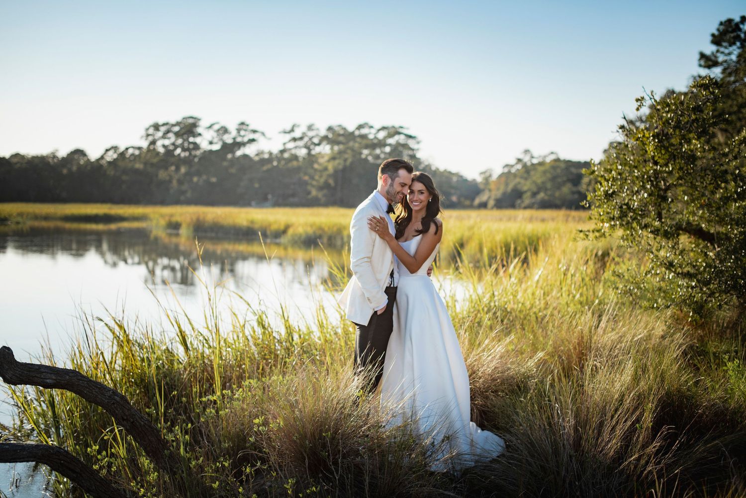 A couple shares a romantic moment in tall grass by a waterfront during golden hour sunset.