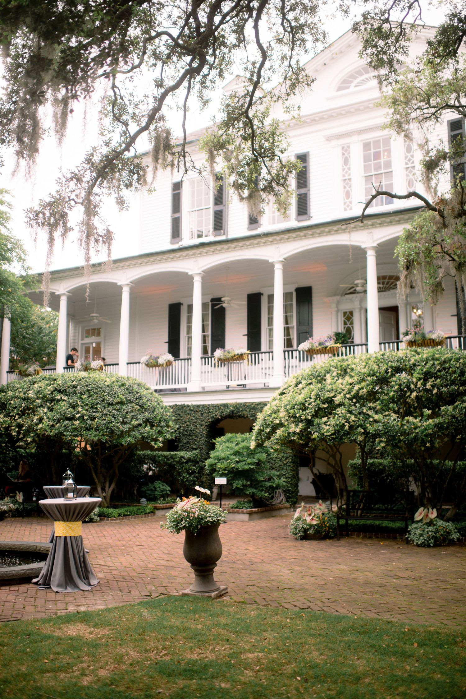 White antebellum mansion with wrap-around columned porch and manicured gardens features brick pathways and ornate planters.
