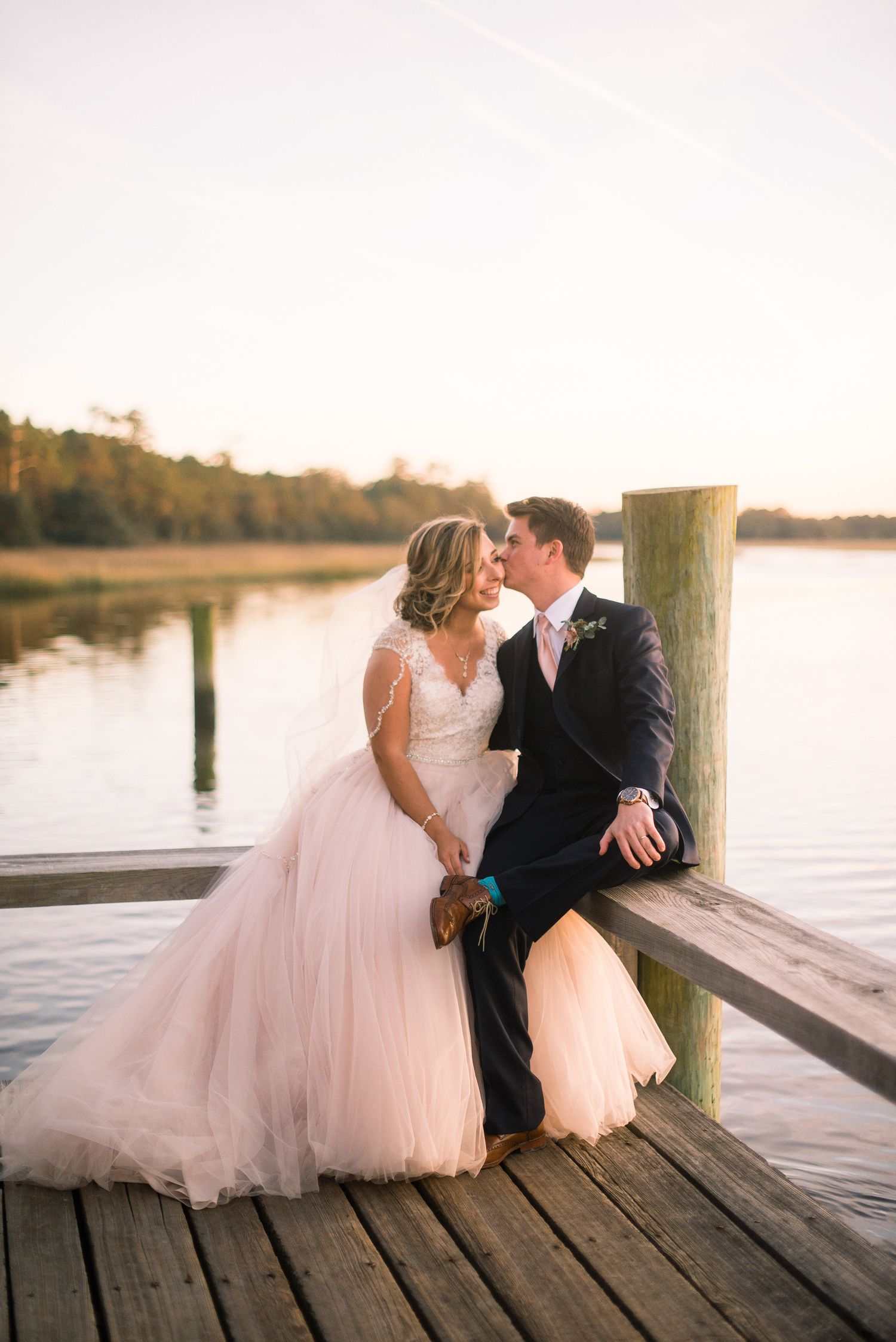 Romantic wedding couple shares intimate moments on a waterfront dock at sunset in a dreamy pink and navy setting.