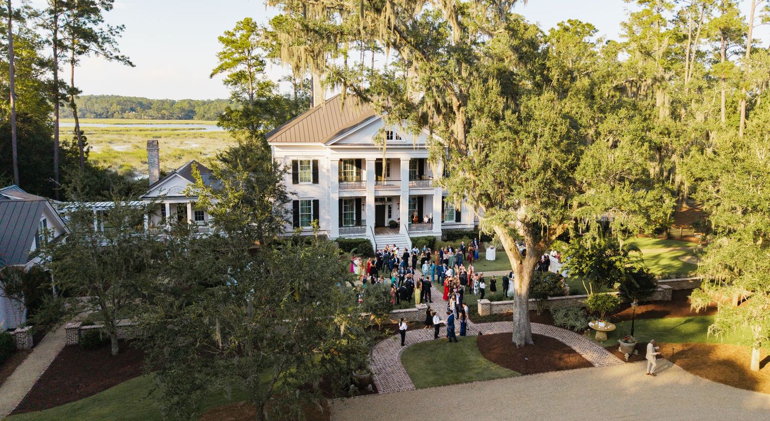 A scenic panoramic view of a historic southern plantation house with white columns and wraparound porches nestled among oak trees.