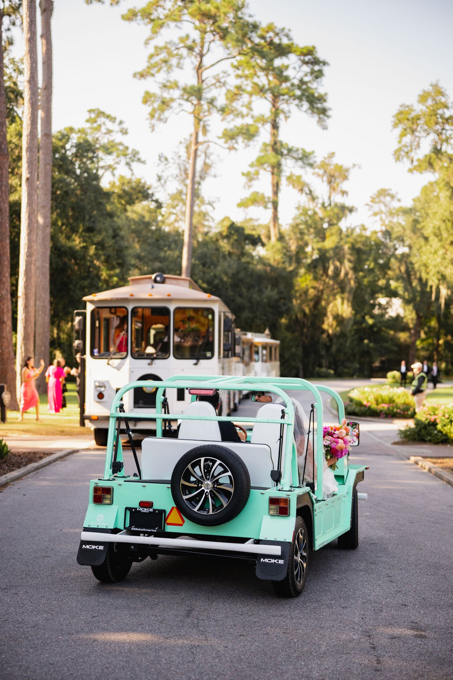 Mint green vintage golf cart follows white trolley on tree-lined resort path at sunset.