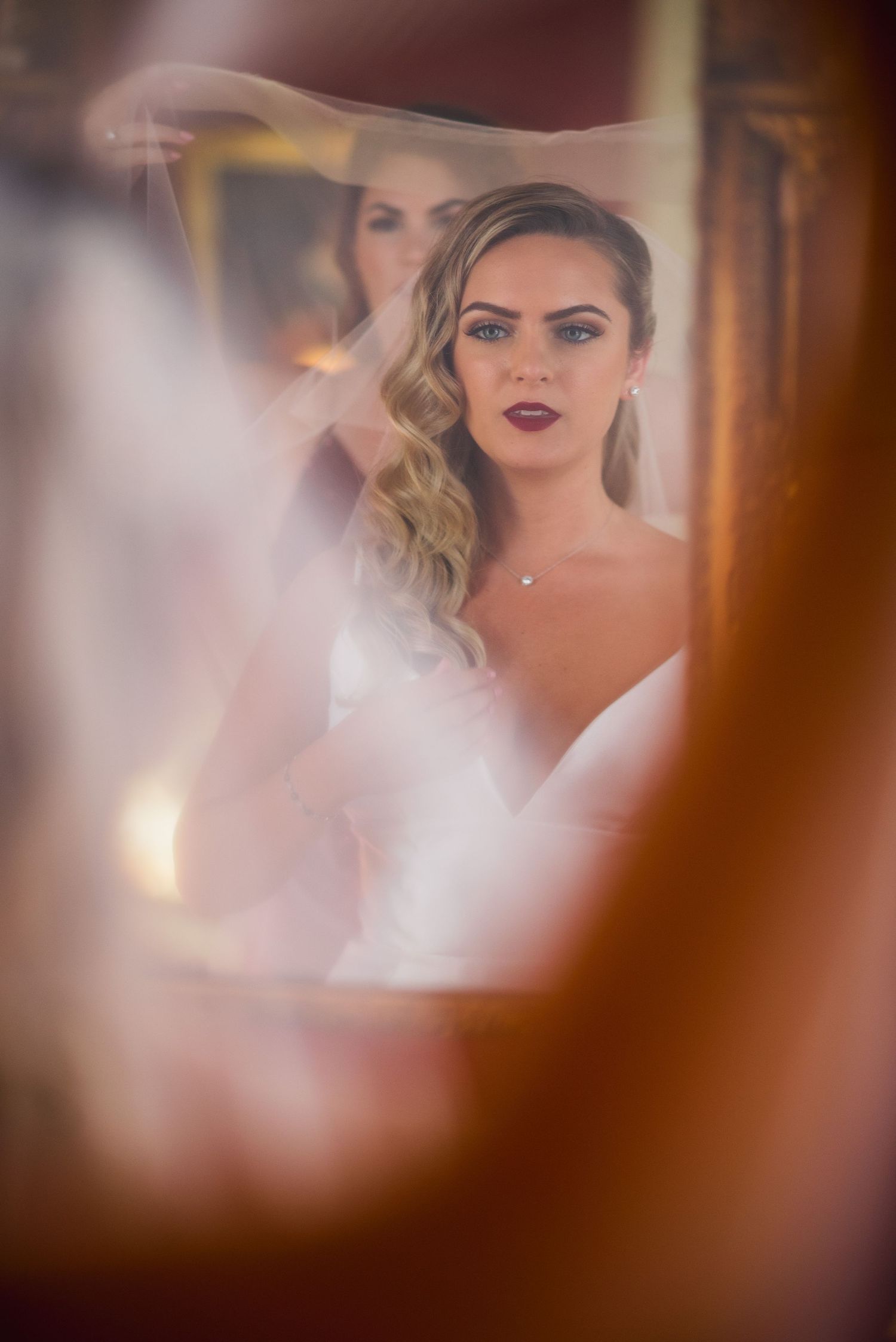 Artistic mirror reflection captures a bride in white with wavy blonde hair and red lipstick on her wedding day.