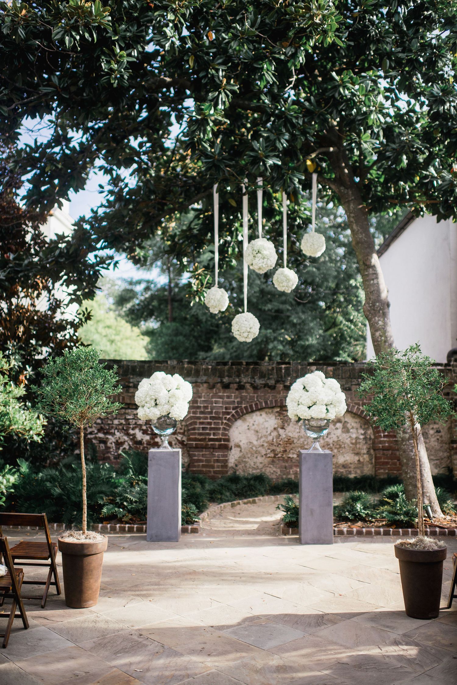 Outdoor wedding ceremony setup with white floral arrangements on pedestals and hanging lanterns under trees by a brick wall.