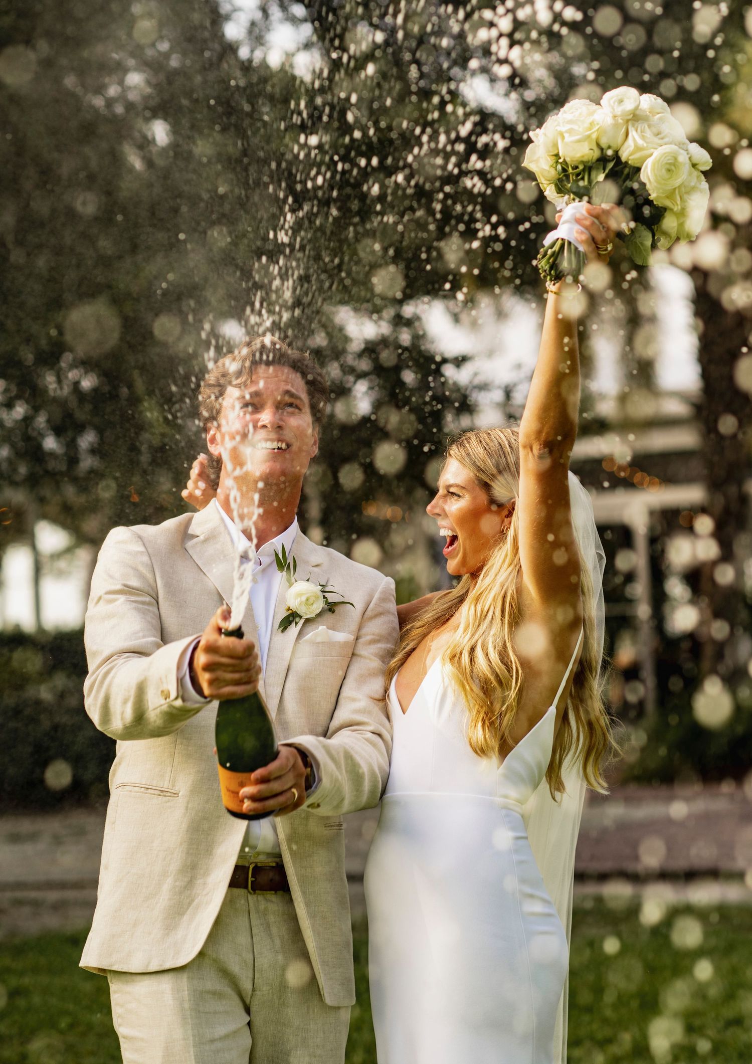 Newlyweds celebrate with champagne spray in an outdoor garden setting wearing white attire.