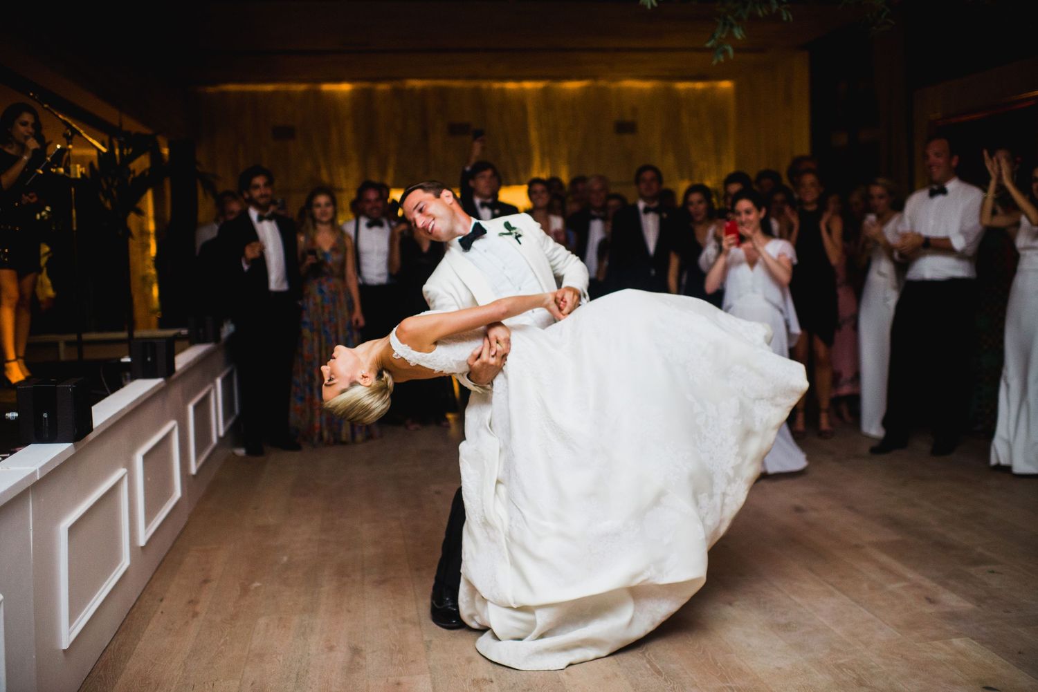 A couple performs their first dance as newlyweds in a dimly lit ballroom surrounded by wedding guests.