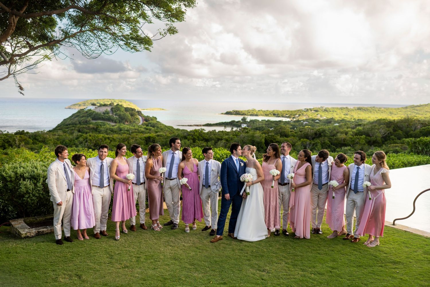 Large wedding party in pink dresses and light suits posed together on a grassy hillside.