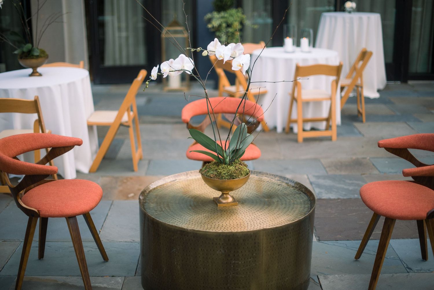 Modern restaurant setup with orange chairs and white tables featuring an orchid centerpiece on a metallic drum table.