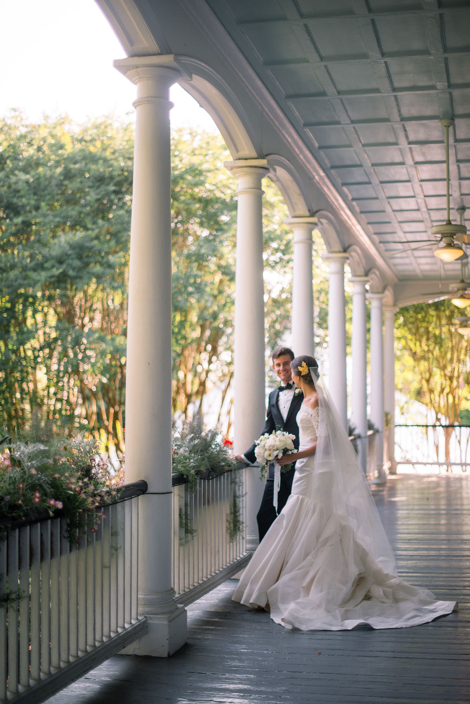 Wedding couple stands beneath a classic white columned porch with natural light filtering through.