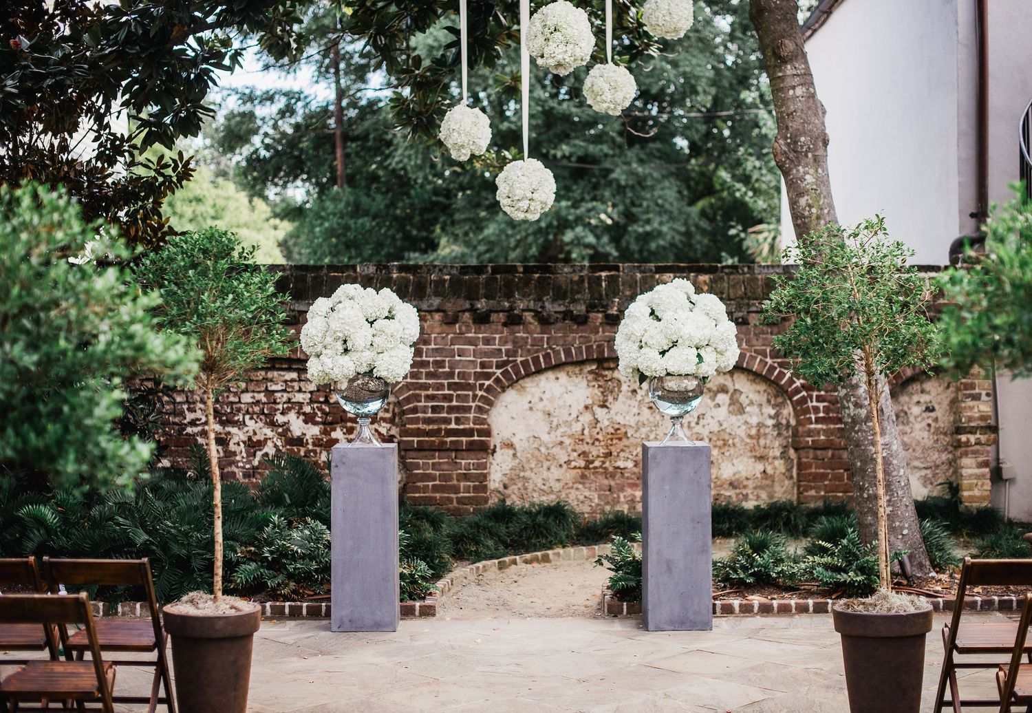 Elegant outdoor garden wedding ceremony setup with white floral arrangements and wooden chairs along a brick wall.