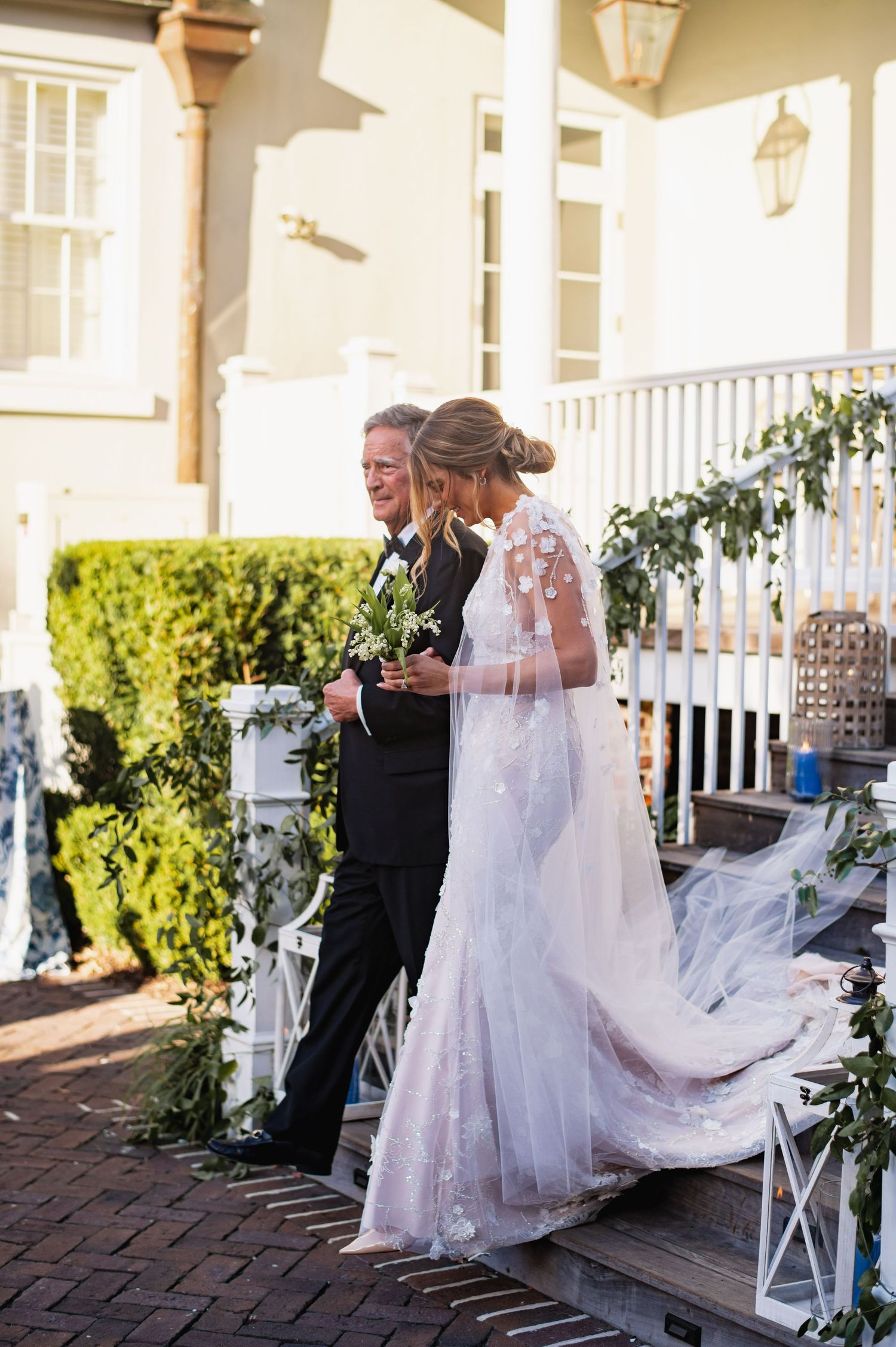 A bride in a lace wedding gown with flowing veil walks down a brick path beside a white porch with lanterns.