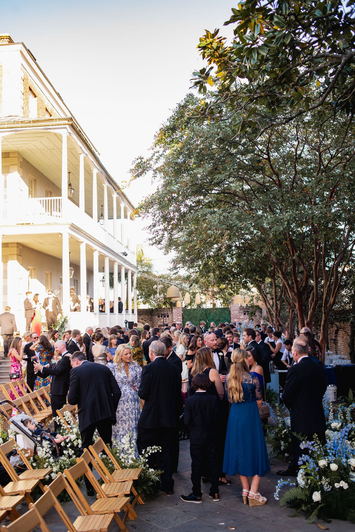 Outdoor wedding reception gathering with guests mingling on a porch decorated with floral arrangements and wooden chairs.