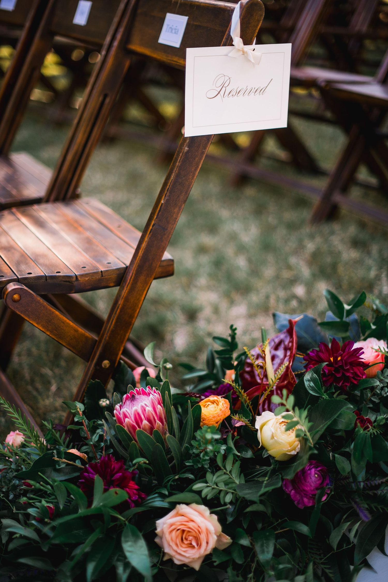 Wooden chairs adorned with fresh floral arrangements featuring pink, orange, and burgundy blooms at an outdoor event.