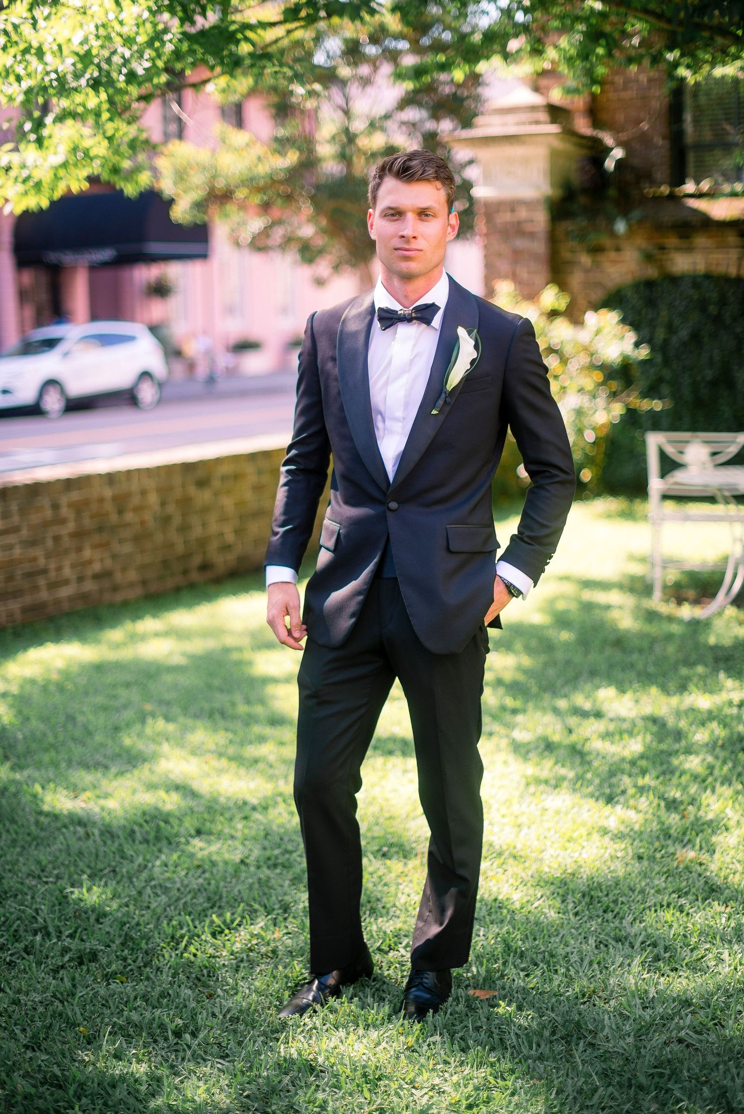 A person in a formal black tuxedo and bow tie stands on grass in a garden setting with sunlit trees in the background.