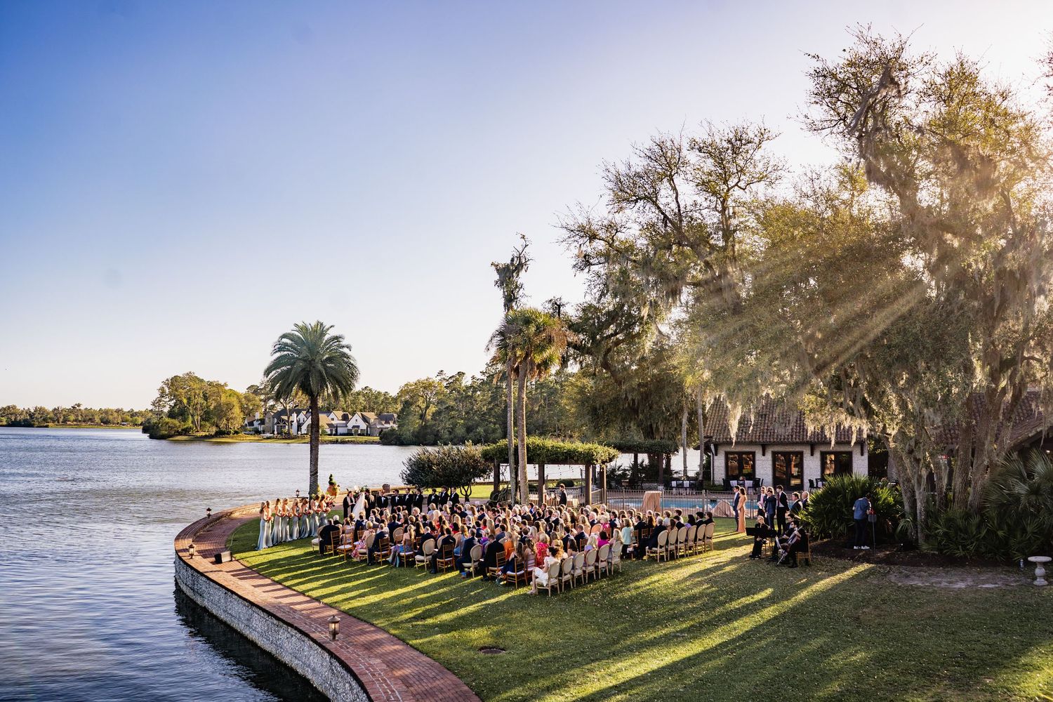 Outdoor wedding ceremony setup on a waterfront lawn with palm trees and sunlight streaming through Spanish moss.