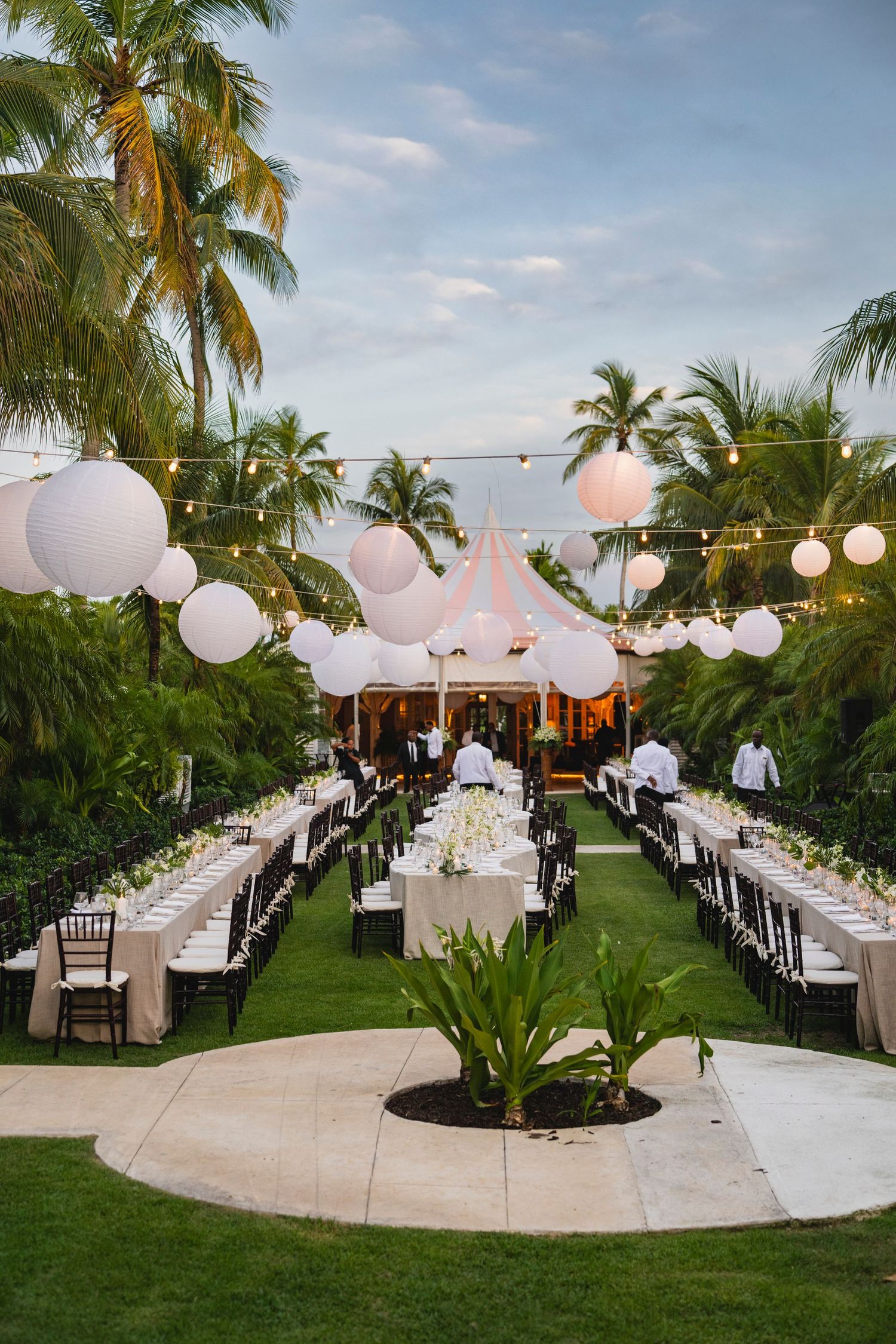 Elegant outdoor wedding reception setup with white lanterns, long tables and palm trees at a tropical beachside venue.