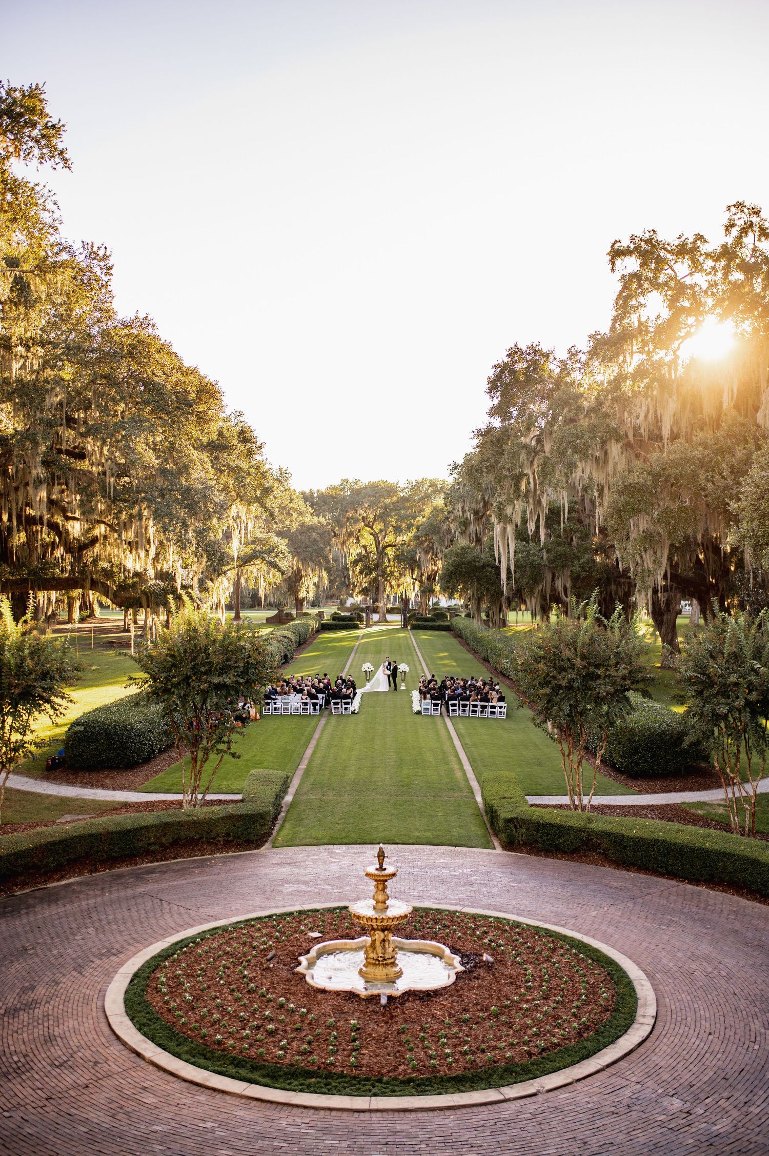Scenic garden pathway with Spanish moss draped trees leads to an ornate fountain at sunset in a formal estate setting.
