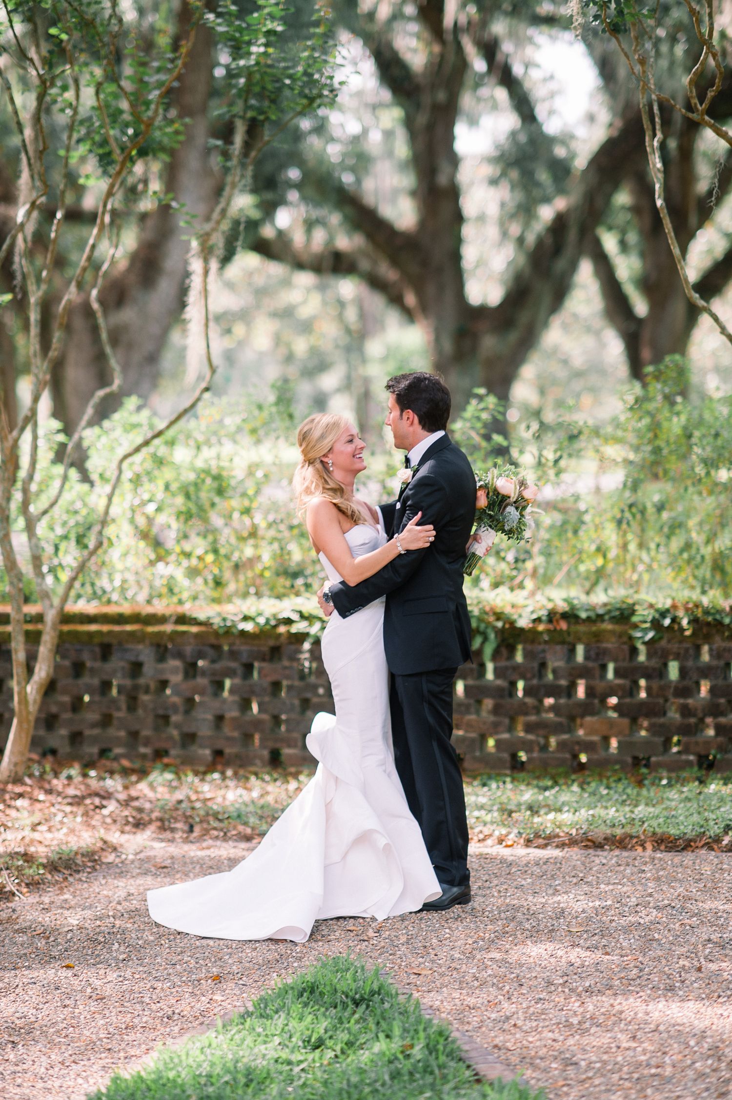 A romantic wedding couple shares an intimate moment on a tree-lined path in a garden setting.