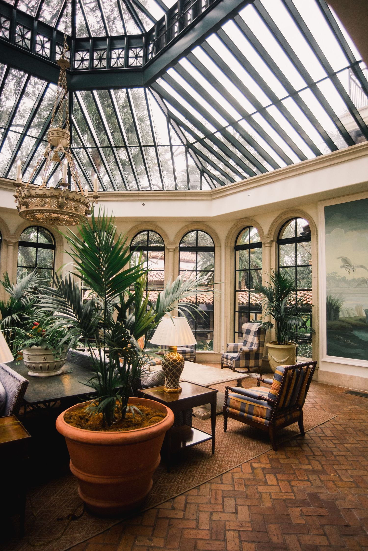 Elegant conservatory sunroom with herringbone brick floors, potted palms, and vintage furniture beneath a glass ceiling.