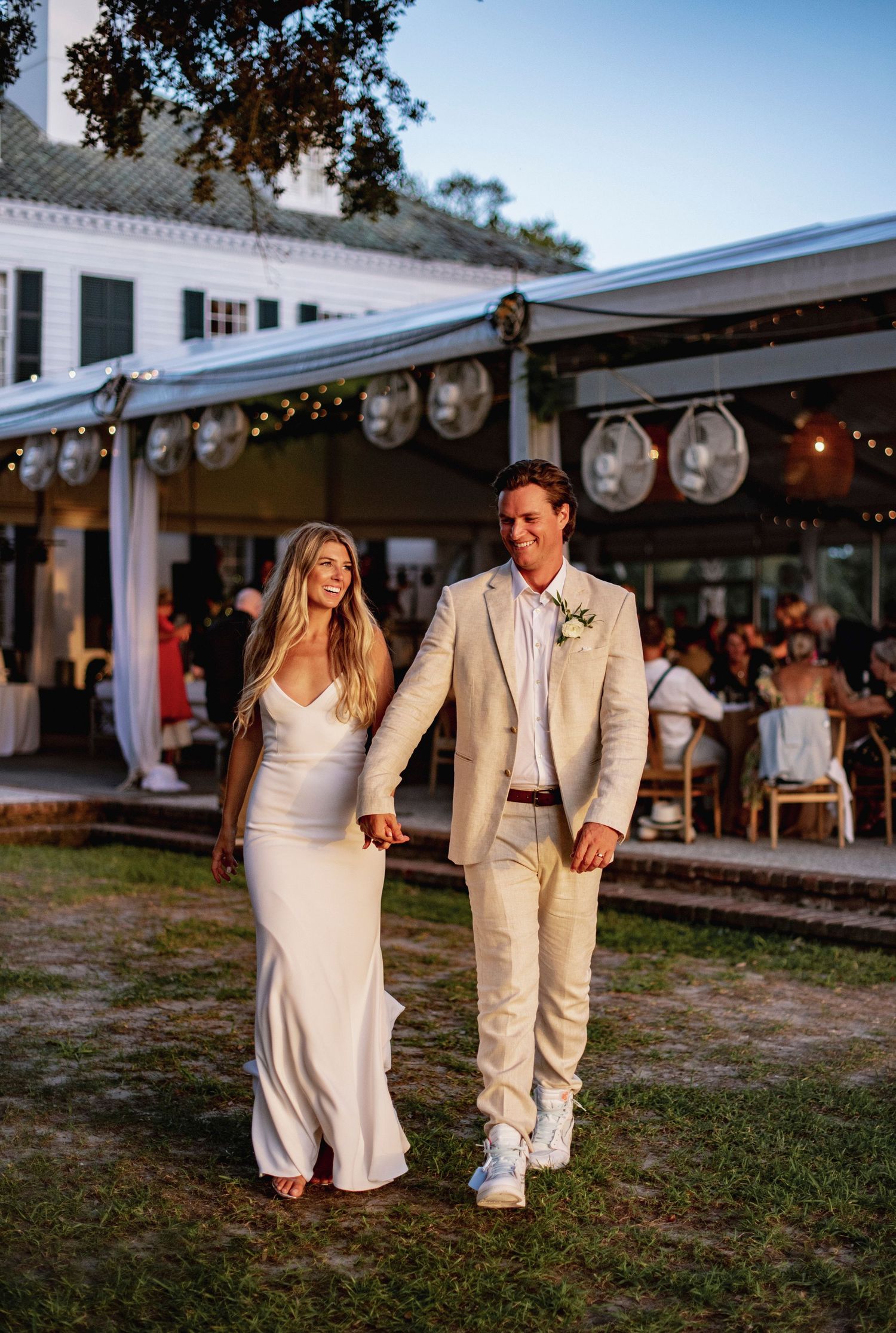 A couple in light-colored wedding attire walk together on grass near a venue decorated with string lights and lanterns.
