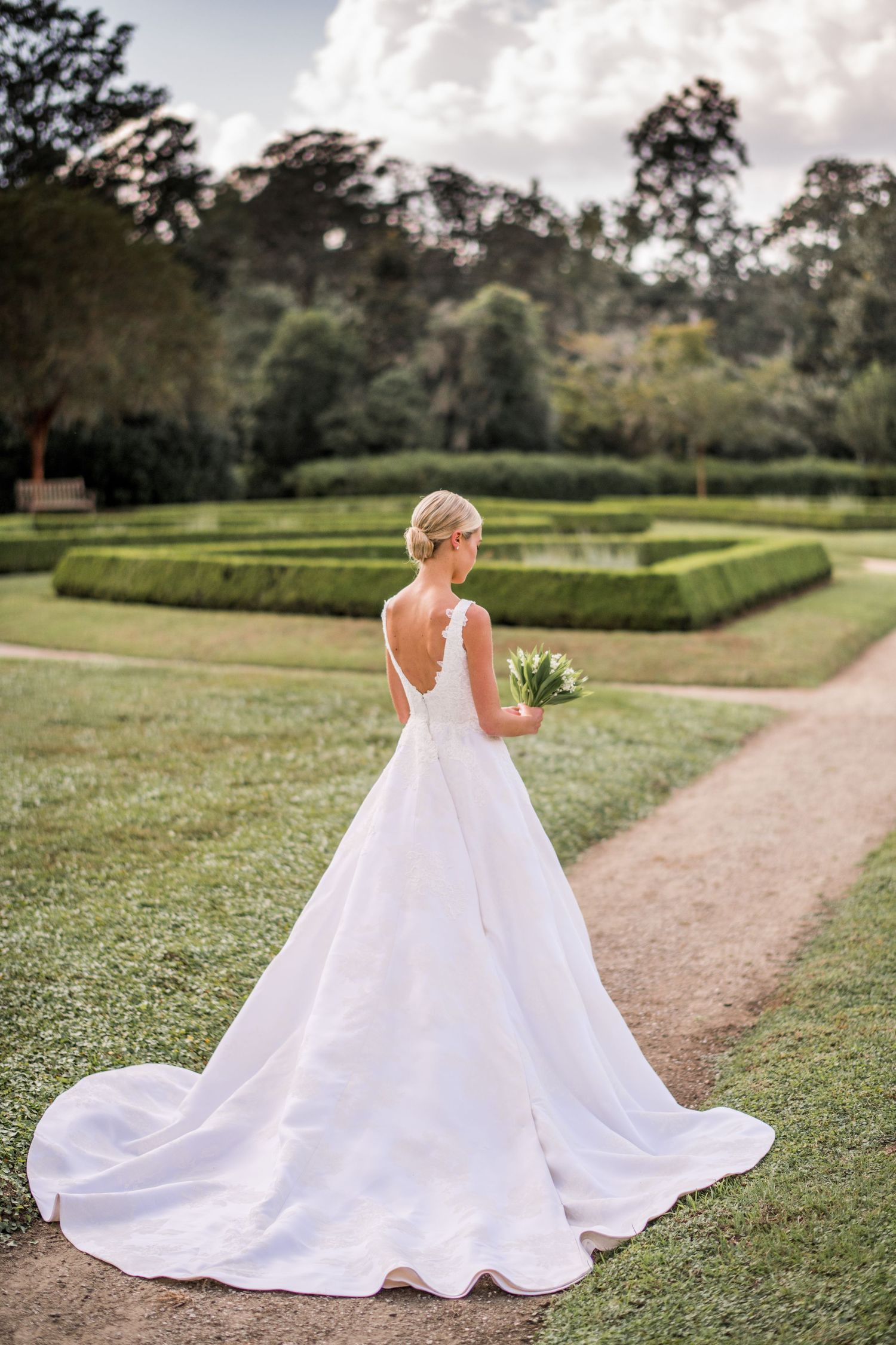 A flowing white wedding dress trails elegantly along a garden path surrounded by manicured hedges and green lawns.