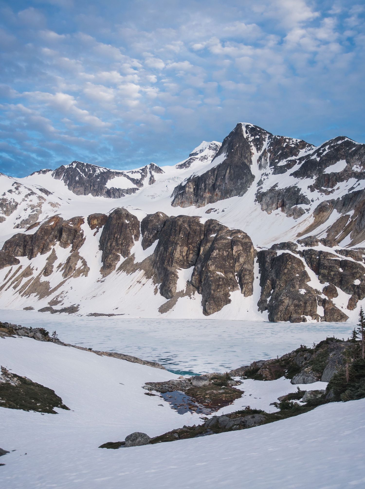 Snow-covered mountain peaks with rugged cliffs and partially frozen alpine lake under scattered clouds.
